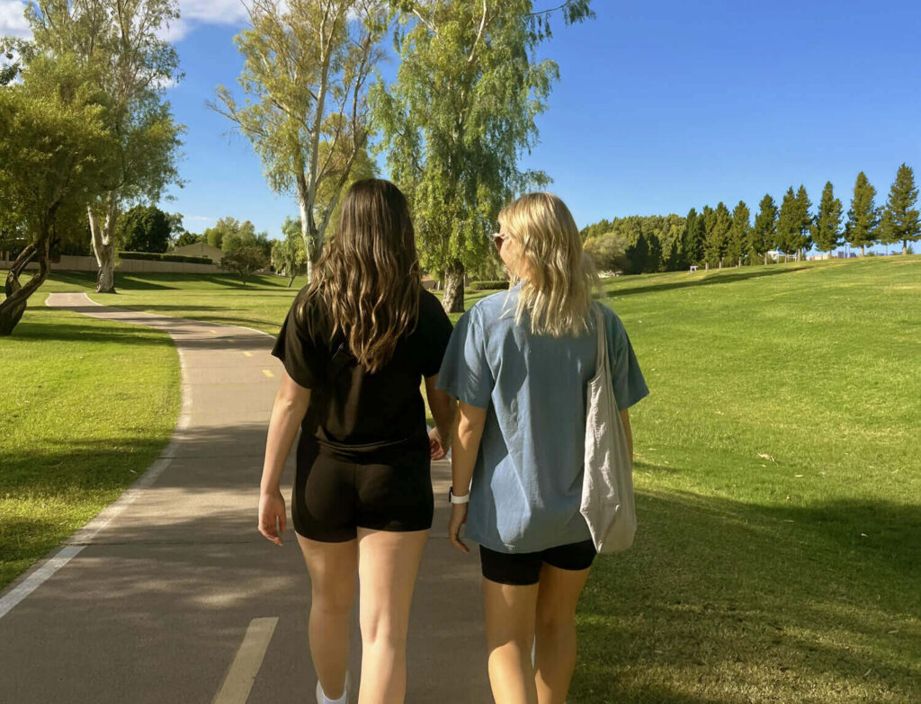Two female friends walking outside along paved path in park e1784039615425
